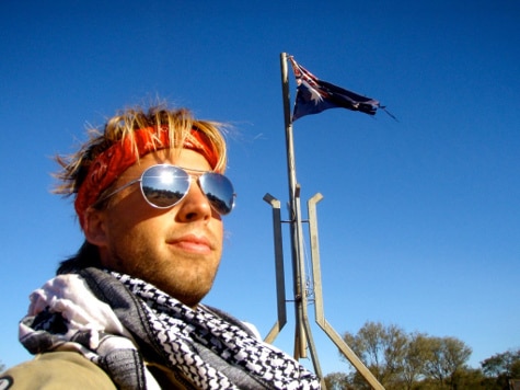 A Motorcycle Expedition Guide confidently poses in front of an Australian flag, sporting sunglasses and a trendy scarf.
