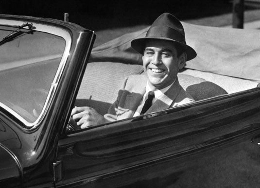 A gentleman in a hat behind the wheel of a convertible car, practicing proper driving etiquette.