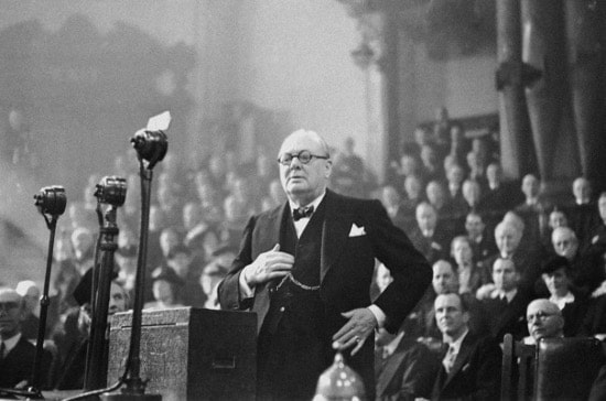 An old photo of a man in a suit standing in front of a crowd, reminiscent of Winston Churchill's public speaking.