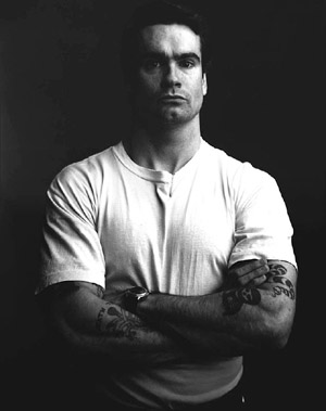 A black and white photo of a man with tattoos, reminiscent of Henry Rollins.